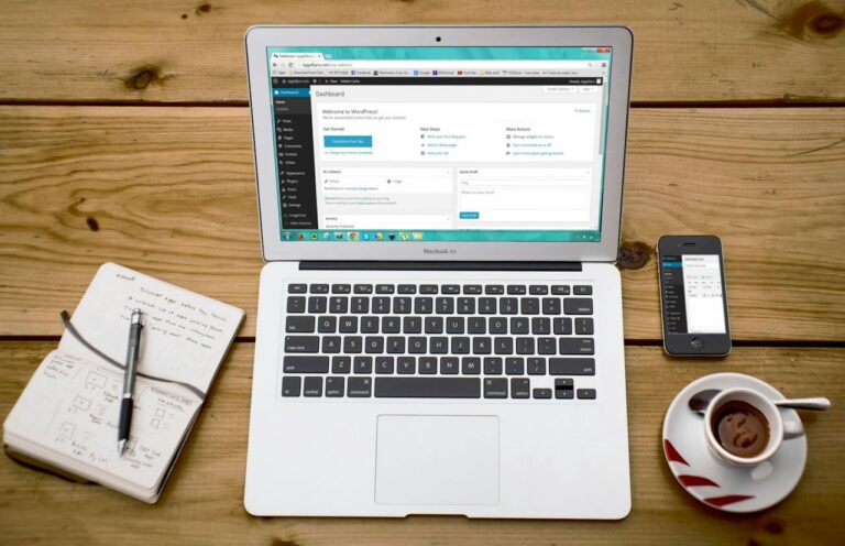 Ein geöffneter Laptop auf einem hölzernen Schreibtisch zeigt einen WordPress-Administrationsbildschirm; daneben liegen ein Notizbuch mit Stift, ein Smartphone und eine Tasse Kaffee mit Löffel, was auf produktive Arbeit in einer gemütlichen Umgebung hinweist.