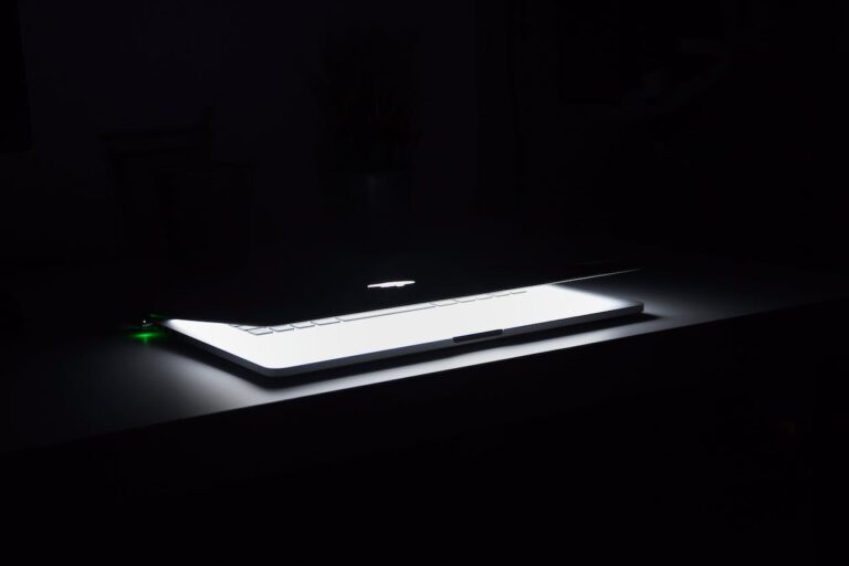 Ein Laptop, wahrscheinlich ein MacBook, ist auf einem dunklen Hintergrund zu sehen, wobei nur die beleuchtete Tastatur und das leuchtende Apple-Logo auf der Rückseite des Bildschirms sichtbar sind.