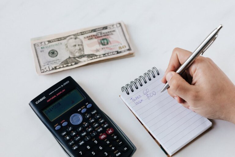 Hand einer Person, die Finanzberechnungen in ein Notizbuch schreibt, neben einem Stapel von 50-Dollar-Scheinen und einem Casio Taschenrechner, was auf Budgetplanung und Finanzmanagement hindeutet.