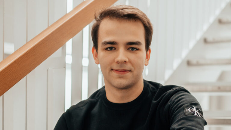 Porträt von Felip Humer-Hager, einem jungen Fachinformatiker für Systemintegration, der lächelnd in die Kamera blickt. Er trägt ein schwarzes T-Shirt und lehnt entspannt an einem Treppengeländer in einer hellen, modernen Umgebung, was Professionalität und Zugänglichkeit ausstrahlt.