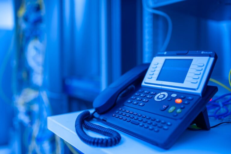 Ein Festnetztelefon mit einem großen Display und zahlreichen Tasten steht auf einem Tisch im Vordergrund, vor dem unscharfen Hintergrund eines Netzwerkservers mit Kabeln und Blinklichtern in einem blau getönten Serverraum, was eine moderne Kommunikationstechnologie in einem IT-Umfeld suggeriert.