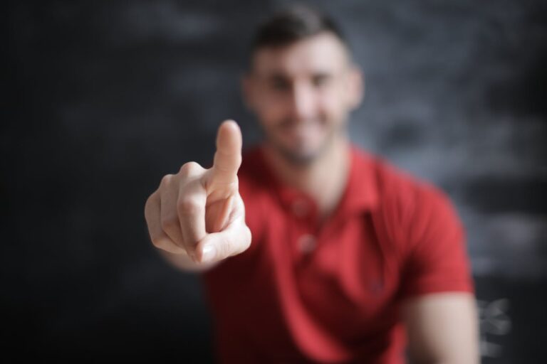Ein unscharf im Hintergrund lächelnder Mann in einem roten Poloshirt streckt seinen Arm aus und zeigt mit dem Finger direkt in die Kamera, was ein einladendes 'Wir suchen dich' Gestik darstellt.