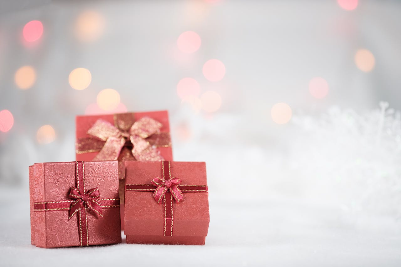 Drei rote Geschenkboxen mit glänzenden goldfarbenen Schleifen auf einer verschneiten Unterlage, im Hintergrund unscharf leuchtende Lichter, die eine festliche Atmosphäre erzeugen. Symbolisch für die freudenreiche Weihnachtszeit und die besten Wünsche von fomedia für das neue Jahr.