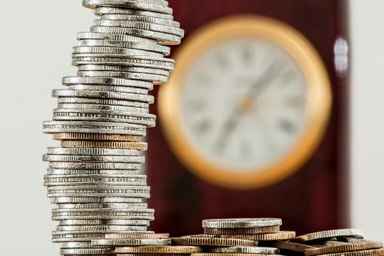 Stapel von Münzen im Vordergrund mit einem unscharfen Hintergrund, auf dem eine Uhr zu erkennen ist, symbolisieren bevorstehende Preisanpassungen, die mit der nächsten Rechnungsstellung wirksam werden.